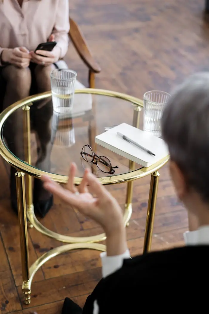hero-img-02 Two adults engaged in a therapy session, seated at a glass table with notebooks and water glasses.